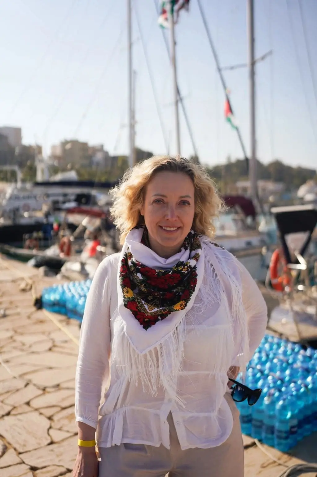 Smiling woman in white blouse and floral scarf stands at a marina with boats in the background on a sunny day.