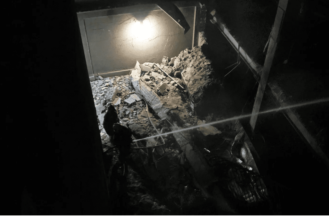 Nighttime construction/ruins scene with two workers amid rubble and debris in a partially collapsed building.