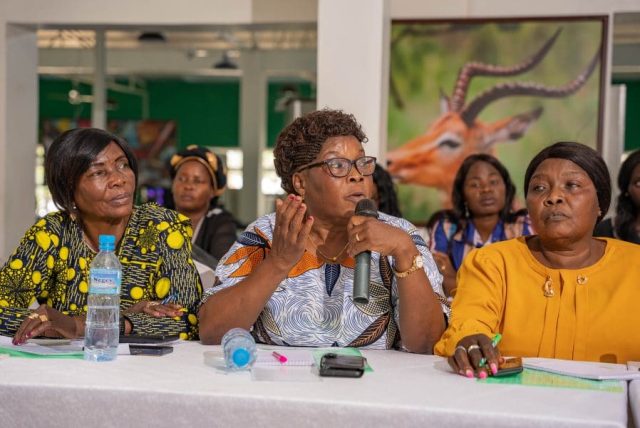 Three women sit in a row, the woman in the centre speaks into a microphone