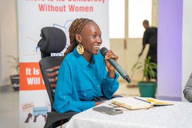 A woman sits at a table, holding a microphone and speaking